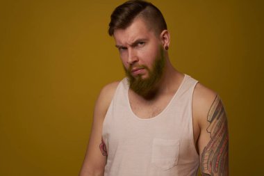 Kollarında dövmeleri olan beyaz tişörtlü sakallı adam ciddi görünüyor.