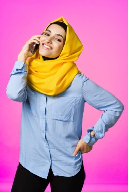 Sarı tesettürlü müslüman kadın telefonda pembe teknolojiyle konuşuyor.