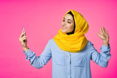 Sarı tesettürlü neşeli bir kadın. Elinde telefon, iletişim teknolojisi var.