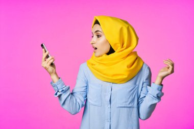 Sarı tesettürlü neşeli bir kadın. Elinde telefon, iletişim teknolojisi var.