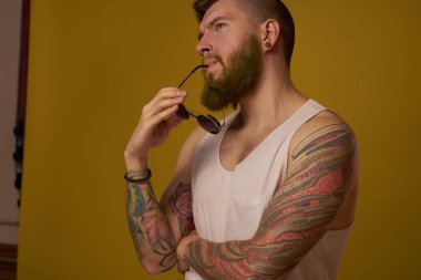 Kollarında beyaz tişörtlü dövme olan bir adam modern moda gözlükleri takıyor.