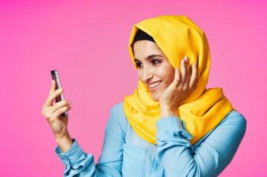 Sarı tesettürlü neşeli bir kadın. Elinde telefon, iletişim teknolojisi var.