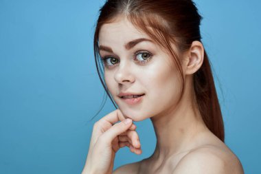 Çıplak omuzlu, at kuyruklu, mavi arka plan pozu veren bir kadın.