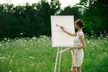 Kadın sanatçı, doğa manzarasında sehpada resim yapar.