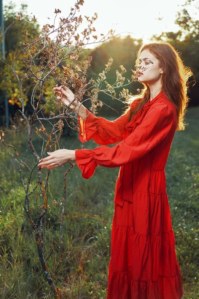 Kvinna i röd klänning i fält nära träd poserar sommar — Stockfoto