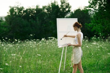 Kadın sanatçı, doğa manzarasında sehpada resim yapar.