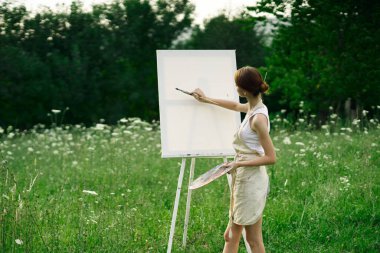 Kadın sanatçı, doğa manzarasında sehpada resim yapar.