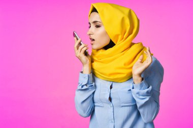 Sarı tesettürlü neşeli bir kadın. Elinde telefon, iletişim teknolojisi var.