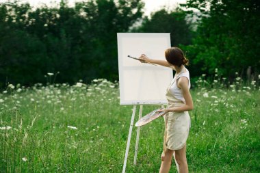 Kadın sanatçı, doğa manzarasında sehpada resim yapar.