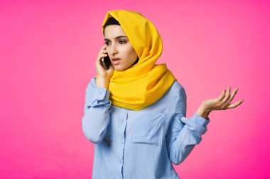 Sarı tesettürlü müslüman kadın telefonda pembe teknolojiyle konuşuyor.