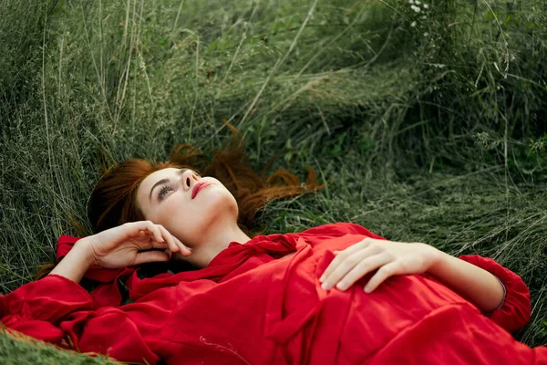 Hezká žena v červených šatech leží na trávě svoboda krajiny — Stock fotografie