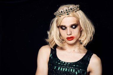 Kadın peruğu giymiş bir adam makyajlı. Kafa travestisi tacını takmış.