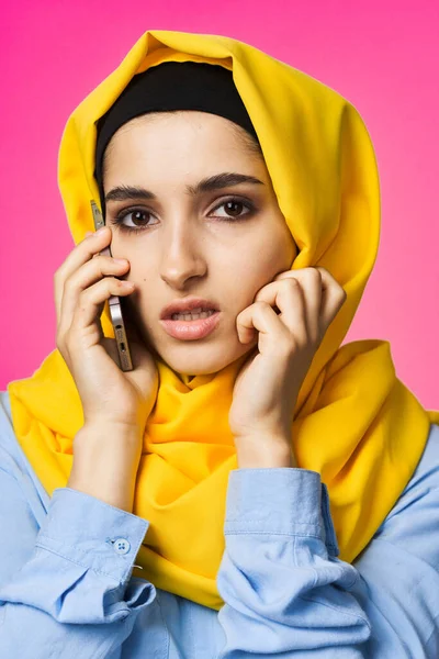 穿着黄色头巾的穆斯林妇女在电话技术上说话粉色背景 — 图库照片