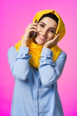 Sarı tesettürlü müslüman kadın telefonda pembe teknolojiyle konuşuyor.