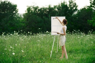 Kadın sanatçı, doğa manzarasında sehpada resim yapar.
