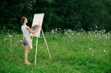 Önlüklü kadın ressam palet boyası doğayı resmediyor.