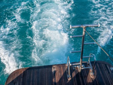 Teknenin kıç tarafında demir bir merdiven. Teknenin arkasındaki suda köpüklü bir patikanın arka planına karşı..