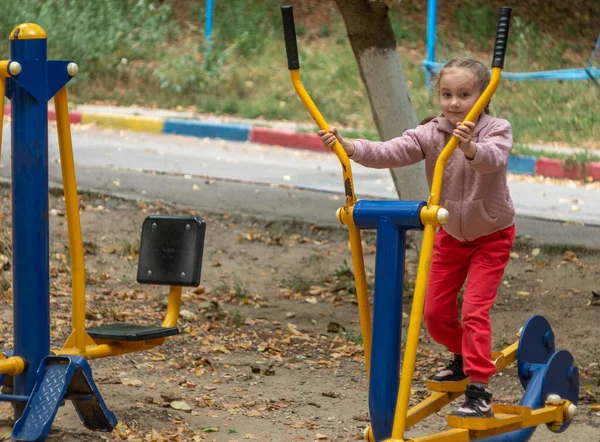 stock image Outdoor sports ground with exercise equipment. A little girl 5-6 years old trains her body muscles on a simulator.