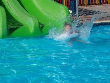 Küçük bir kız, su parkındaki mavi berrak suyla yeşil bir su kaydırağından aşağı yuvarlanıyor. Tatil köyünde yaz tatili.