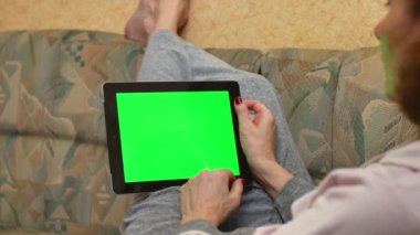 An adult woman holding a tablet computer with a green screen lies on the sofa at home. View from the back. Online learning and work. Chroma key screen.