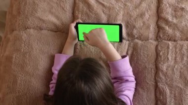 Yatakta oturan 7-8 yaşlarında küçük bir kız önceden ayarlanmış yeşil ekranlı akıllı telefon kullanıyor. Ekran düzeni için ideal. Krom anahtar. Yeşil Ekran.