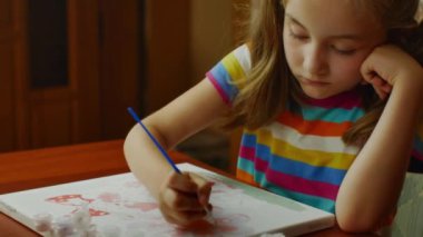 The girl sitting at the table enthusiastically draws with a brush and watercolor paints on a white canvas of paper. Drawing lessons for children and teenagers.