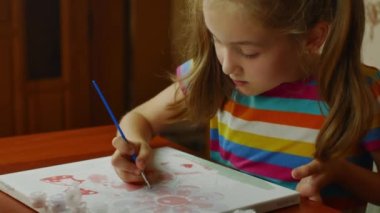 The girl sitting at the table enthusiastically draws with a brush and watercolor paints on a white canvas of paper. Drawing lessons for children and teenagers.