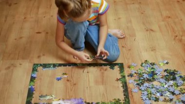 A pretty girl dressed in a gray jumper and blue jeans with interest collects puzzles on a wooden floor. Children playing.