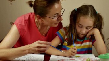 A young beautiful grandmother teaches her pretty granddaughter to paint with paints. Home education of children and adolescents in the drawing.