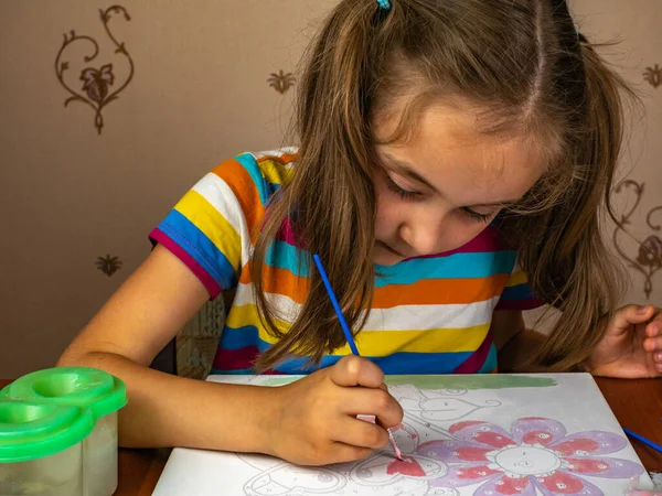 The girl sitting at the table enthusiastically draws with a brush and watercolor paints on a white canvas of paper. Drawing lessons for children and teenagers.