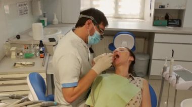 Dentists office. The patient lies in the dental chair with her mouth open, while the dentist sets up the dental bur machine for drilling teeth. 
