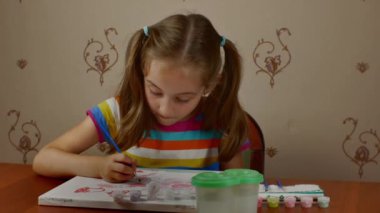 A pretty girl of 7-8 years old paints with paints on a white canvas sitting at a table. Childrens creativity. Teaching young people the art of painting.