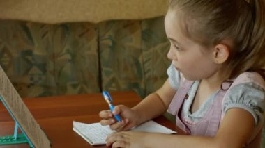 A young pretty schoolgirl is doing her homework while sitting at the dining table at home. Primary school education.