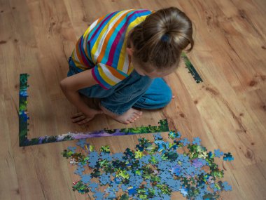 A smart cute girl in a multi-colored t-shirt and blue jeans assembles a complex puzzle consisting of small pieces on a wooden floor close-up. High-quality 4k footage.