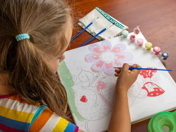 A little girls hand drawing with a brush and watercolor paints on white paper. Close-up. View from above. Creativity develops children.