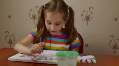 A pretty girl of 7-8 years old paints with paints on a white canvas sitting at a table. Childrens creativity. Teaching young people the art of painting.