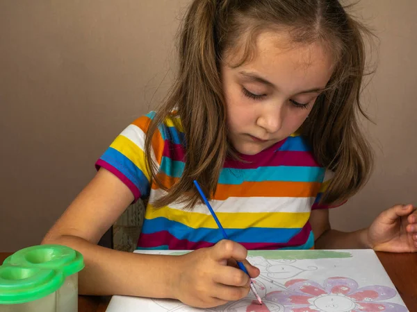 A cute little girl sits at a table and draws with paints on white paper. A beautiful little girl draws with pleasure while sitting at the table. Close-up. The concept of teaching children to draw