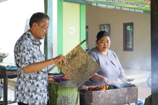 Şişman kadın yaşlı adamın satay pişirmesine yardım ediyor.