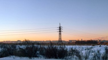 Bir elektrik hattı direği, akşam gökyüzünün ve şehrin arka planında, batan güneşin altında bir kış tarlasında duruyor.