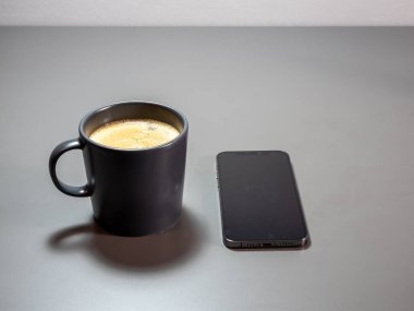 A cup of coffee and a phone with a black screen on a gray table. Coffee and telephone on a gray table.