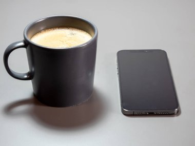 A cup of coffee and a phone with a black screen on a gray table. Coffee and telephone on a gray table.