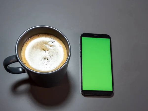 A cup of coffee and a phone with a green screen on a gray table. Coffee and telephone on a gray table.