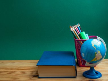 A book, a metal cup with pencils and a globe on the table against the background of a green board. School.