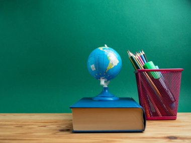 A book, a metal cup with pencils and a globe on the table against the background of a green board. School.
