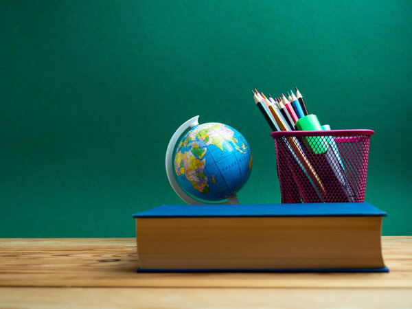 A book, a metal cup with pencils and a globe on the table against the background of a green board. School.