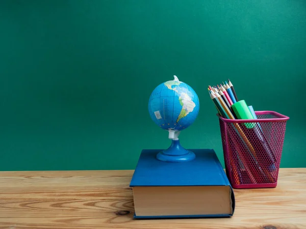 A book, a metal cup with pencils and a globe on the table against the background of a green board. School.