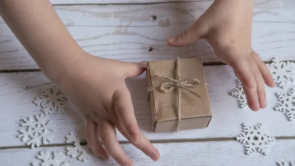 Una mano pequeña de los niños alcanza para un regalo de Navidad embalado en el embalaje del cangrejo en un fondo blanco de madera con los copos de nieve de su árbol —  Fotos de Stock