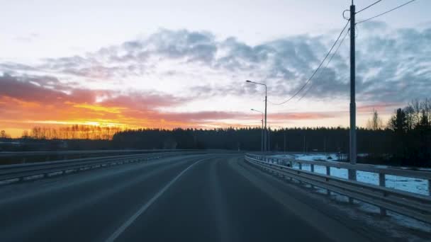 Вид через лобовое стекло движущегося автомобиля на дороге с забором и красивым восходом солнца в зимнем небе. — стоковое видео