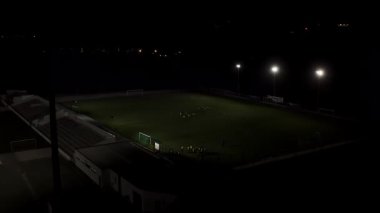 Gece hava manzaralı futbol stadyumu. Futbol sahasında antrenmandaki oyuncularla projektörlü hava aracı uçuşu.. 