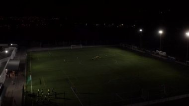 Gece hava manzaralı futbol stadyumu. Futbol sahasında antrenmandaki oyuncularla projektörlü hava aracı uçuşu.. 